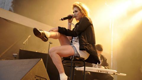 Die US-Sängerin Uffie sitzt während eines Konzertes auf einem Barhocker, singt ins Mikrofon und schlägt die Beine übereinander. Sie wird von hinten von einem gelblichen Licht angestrahlt, im Hintergrund sitzt ein Keyboarder.