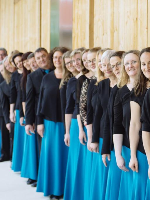Der Chor steht in einer langen Schlange an einer Holzhausfront aufgestellt. Die Damen tragen dabei hellblaue Röcke und Oberteile aus Spitze.