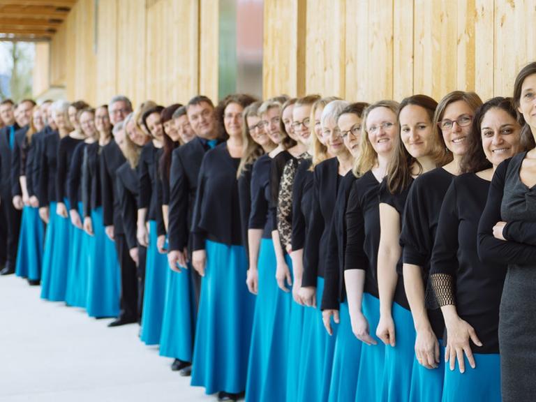 Der Chor steht in einer langen Schlange an einer Holzhausfront aufgestellt. Die Damen tragen dabei hellblaue Röcke und Oberteile aus Spitze.