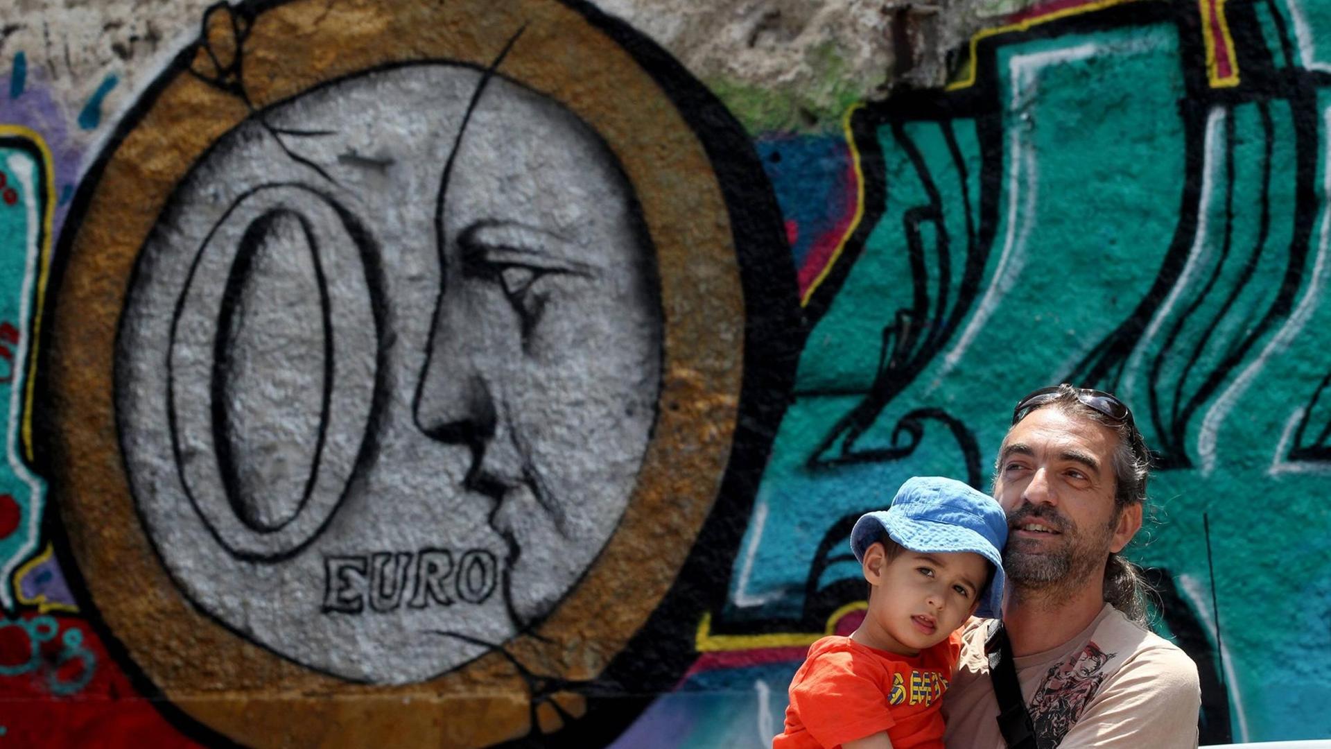 Ein Mann steht mit seinem Kind auf dem Arm vor einem Graffiti in Athen, das eine Euro-Münze zeigt.