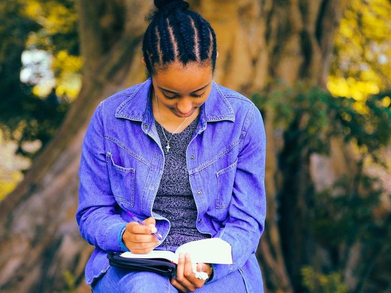 Eine Schwarze Frau sitzt hält einen Stift in der Hand und liest dabei in einem Buch.