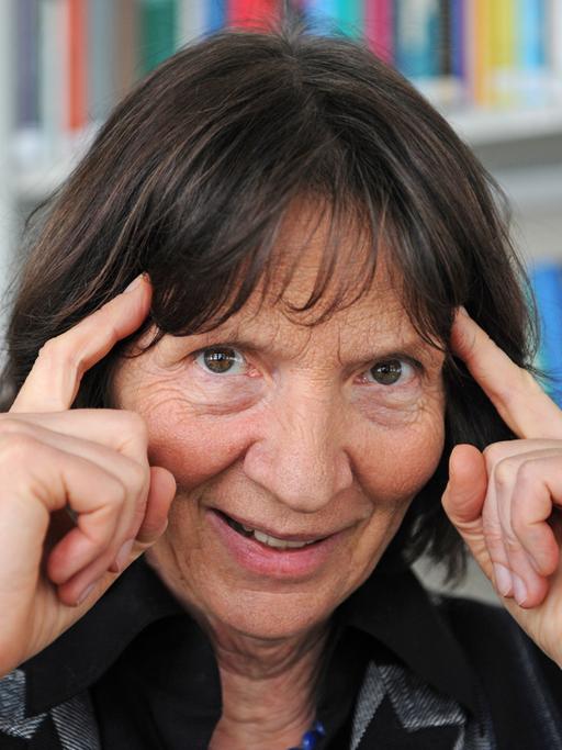 Die Kulturwissenschaftlerin Aleida Assmann, vor einem Bücherregal, hält sich ihre Zeigefingern an die Schläfe.