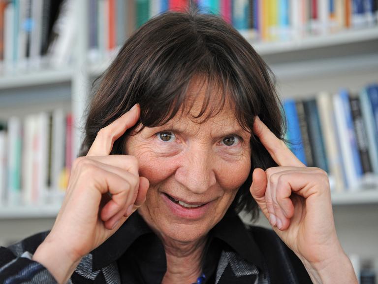 Die Kulturwissenschaftlerin Aleida Assmann, vor einem Bücherregal, hält sich ihre Zeigefingern an die Schläfe.
