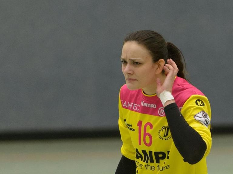 Eine Handballspielerin führt die linke Hand zu ihrem linken Ohr, um etwas zu hören. Sie schaut fragend.