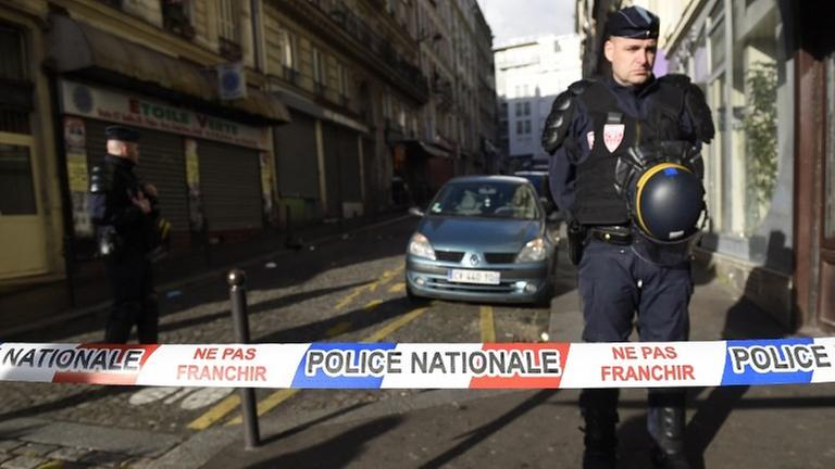 Ein Polizist steht hinter einem rot-weiß-blauen Absperrband mit der Aufschrift "Police Nationale"