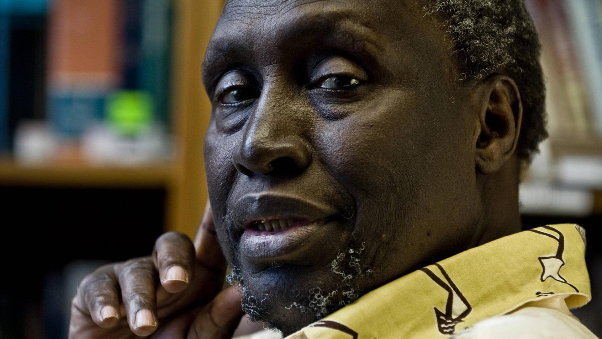 Portrait des kenianischen Schriftstellers Ngugi Wa Thiong o, der nach linke in die Kamera guckt und die rechte Hand mit Daumen und Zeigefinger an die Wange hält.