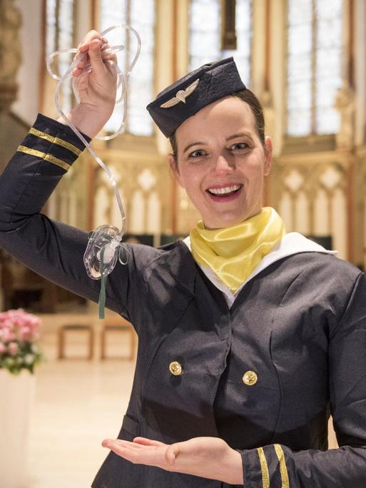 Eine Stewardess steht in einem Kirchenschiff und hält eine Sauerstoffmaskein die Höhe.
