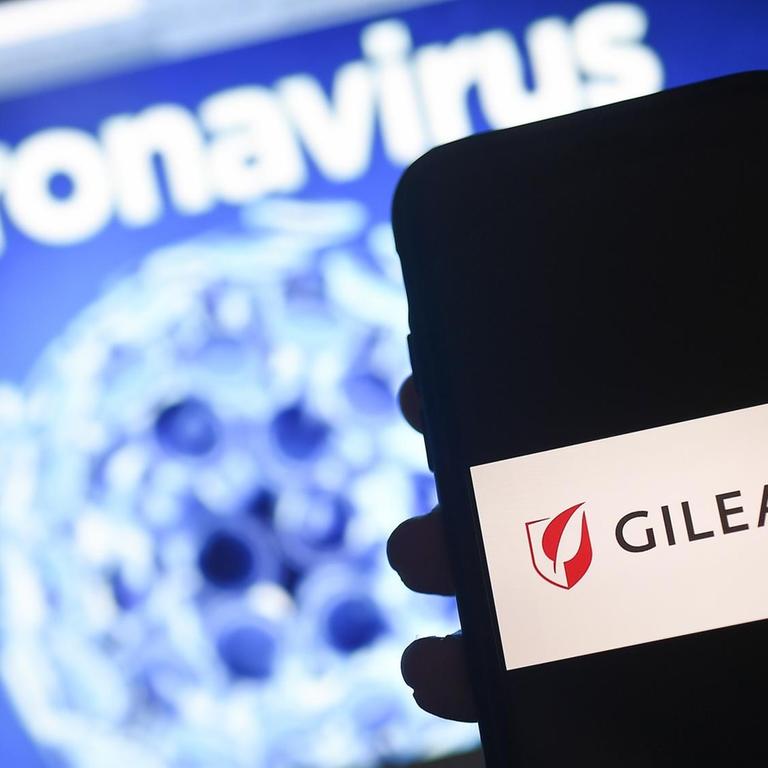 Das Gilead-Logo wird auf einem Smartphone neben einem Bildschirm mit einer Coronavirus-Grafik