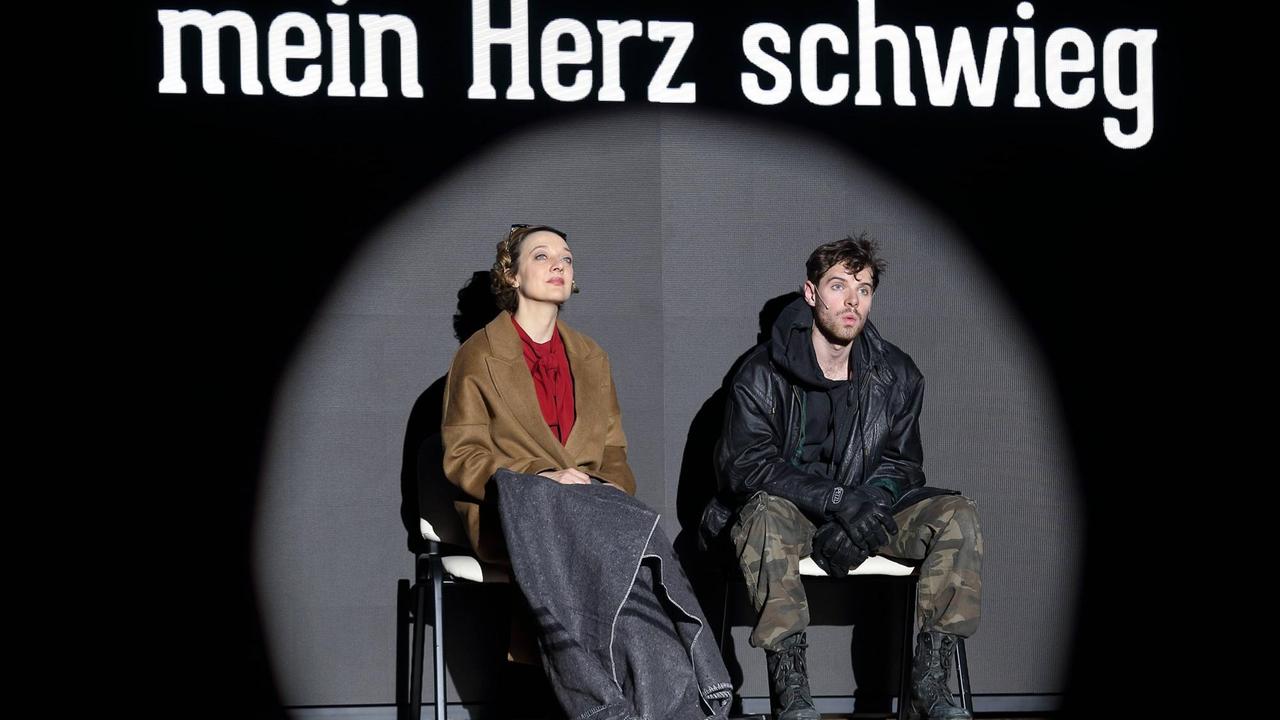 Foto von der Inszenierung: Ein Mann und eine Frau sitzen auf Stühlen auf der Bühne. Über ihren Köpfen ist der Schriftzug "Mein Herz schwieg" zu lesen.