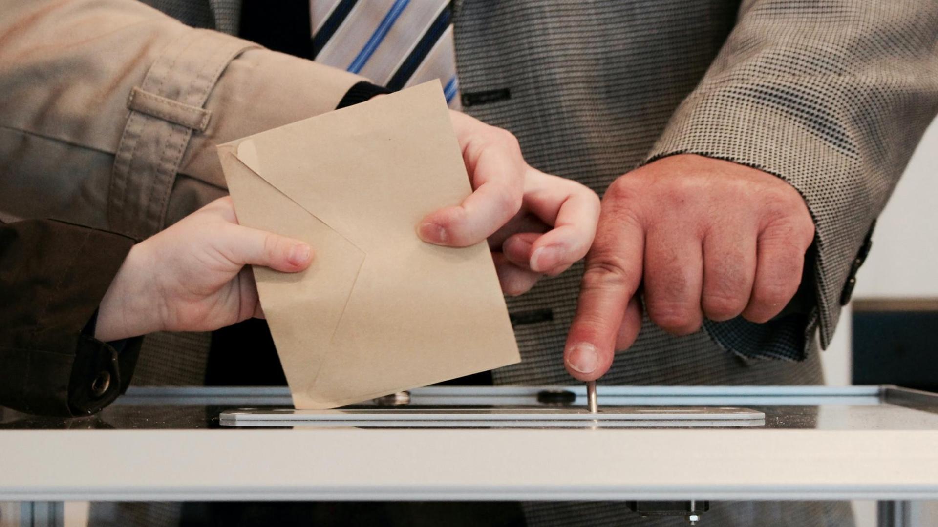 Ein Wahlbriefumschlag und die Hand eines Kindes, einer Frau und eines Mannes.