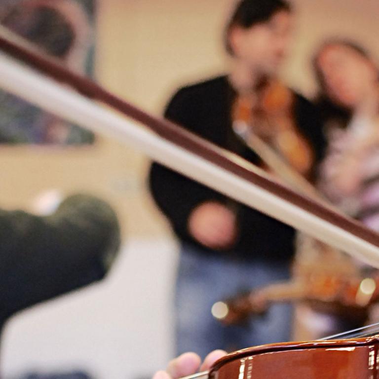 Musikunterricht an einer Waldorfschule: Im Vordergrund spielt eine Person Geige, im Hintergrund sind unscharf zwei Schülerinnen beim Geigespielen zu sehen.