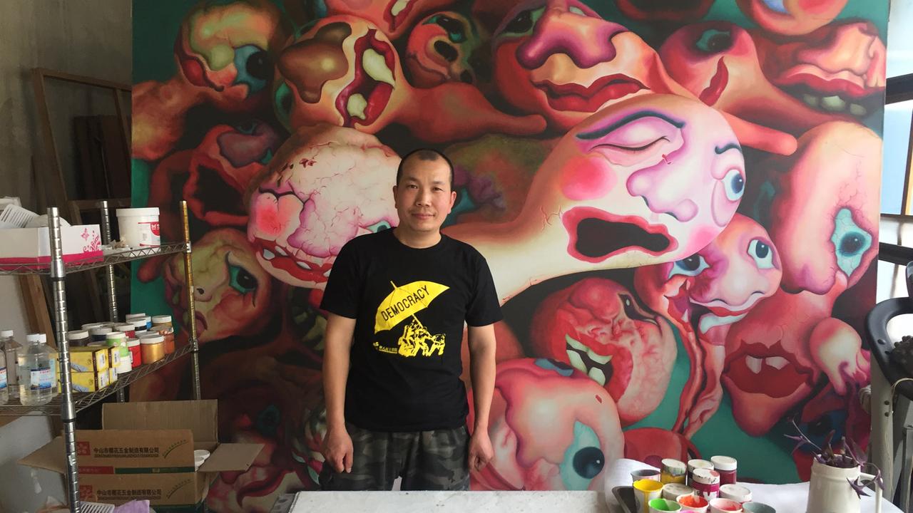 Der chinesische Künstler Lv Shang steht in seinem Atelier vor einem seiner Bilder. Es sind verzerrte Köpfe und Gesichter zu sehen. 