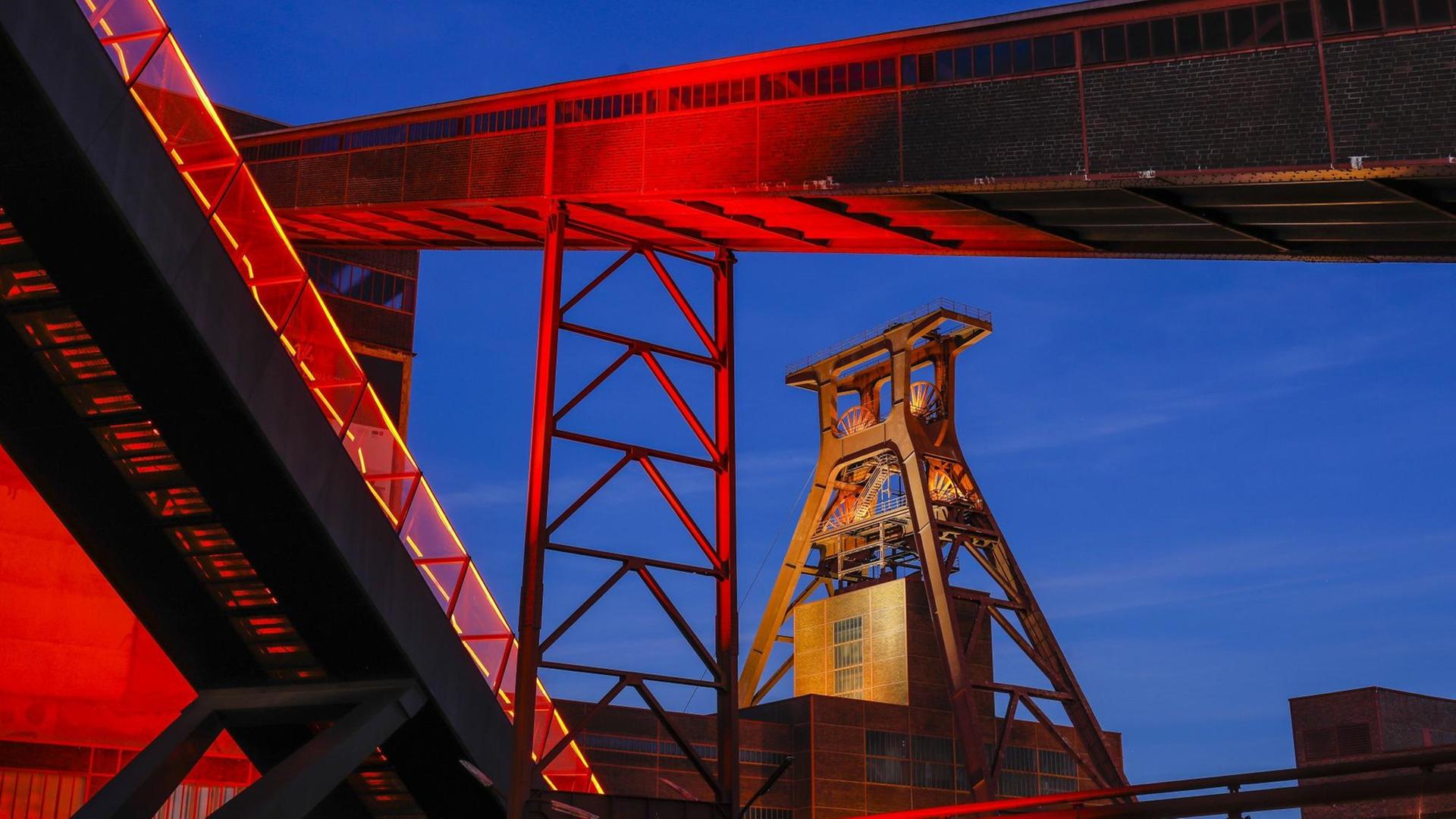 Das Foto zeigt die Zeche Zollverein in Essen. Der über das Ruhrgebiet hinaus bekannte Ausstellungs- und Veranstaltungsort ist in der Abenddämmerung rot illuminiert.