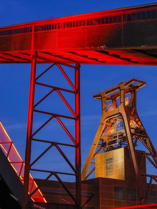 Das Foto zeigt die Zeche Zollverein in Essen. Der über das Ruhrgebiet hinaus bekannte Ausstellungs- und Veranstaltungsort ist in der Abenddämmerung rot illuminiert.