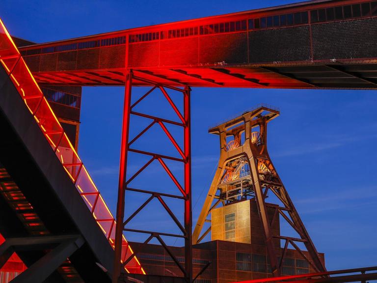 Das Foto zeigt die Zeche Zollverein in Essen. Der über das Ruhrgebiet hinaus bekannte Ausstellungs- und Veranstaltungsort ist in der Abenddämmerung rot illuminiert.