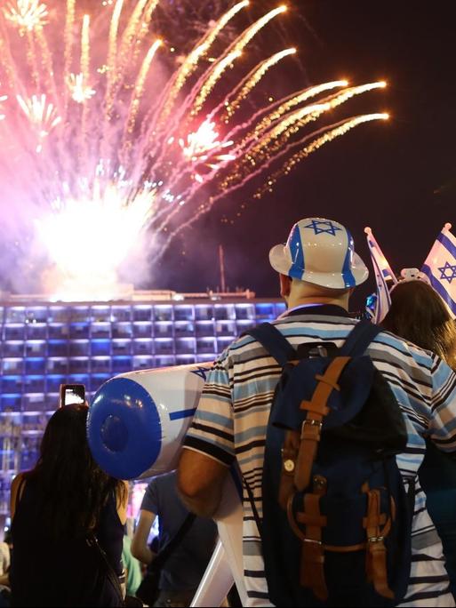 Das Bild zeigt Menschen auf dem Rabin-Platz in Tel Aviv. Sie schauen ein Feuerwerk an, einer trägt einen Hut in den Farben der Israelflagge.
