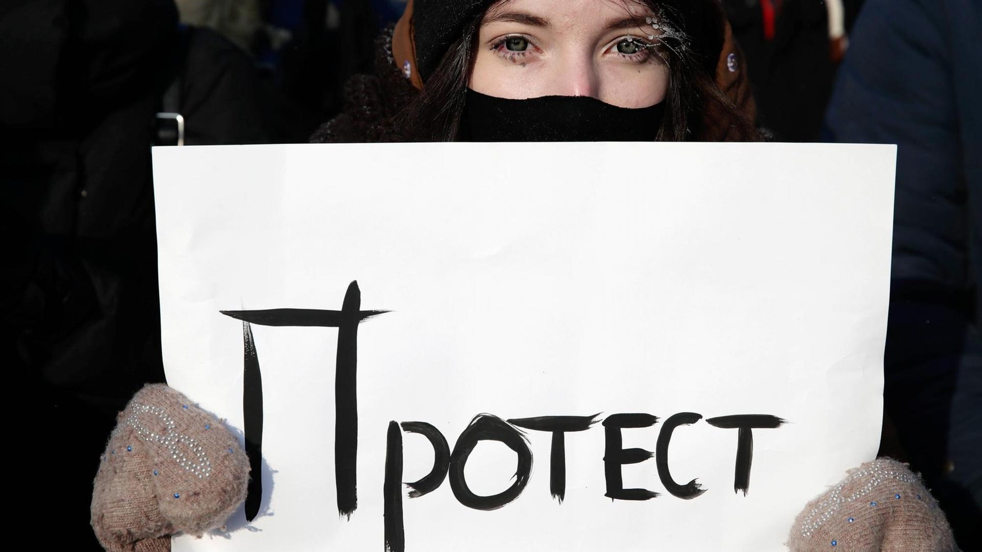 Eine junge Frau mit Wollhandschuhen, Mütze und Mundschutz hält ein Schilf, auf dem in kyrillischer Schrift "Protest" steht.