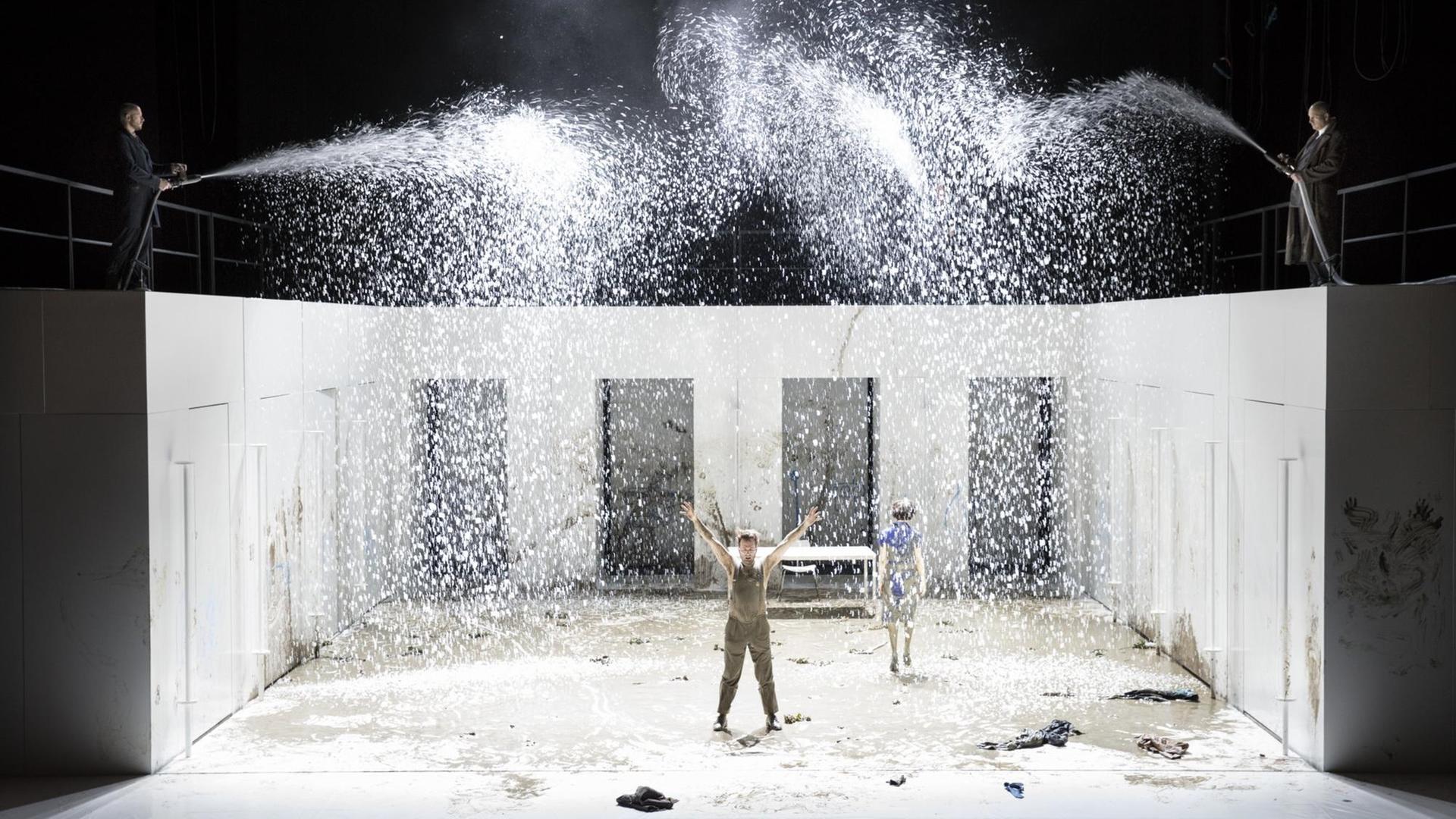 Szene aus "Les Bienveillantes": Zwei Schauspieler sprenkeln mit Schläuchen Wasser auf die völlig verdreckte Bühne und die darauf stehenden Schauspieler.