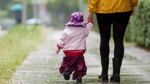 Ein kleines Mädchen läuft mit Regenkleidung an der Hand ihrer Mutter über einen Gehweg