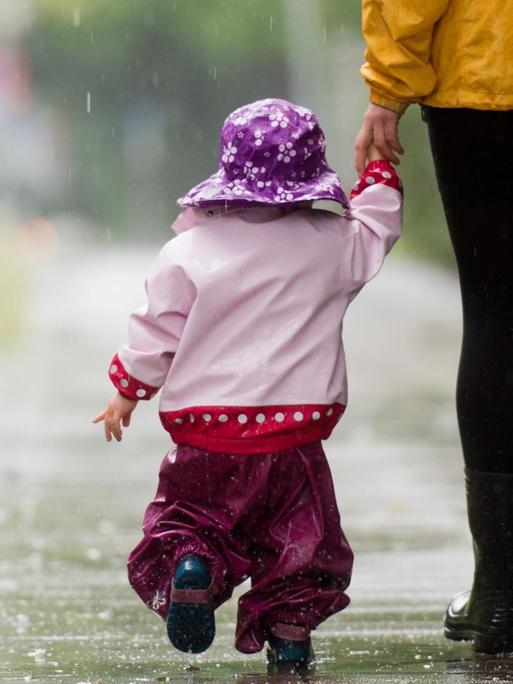 Ein kleines Mädchen läuft mit Regenkleidung an der Hand ihrer Mutter über einen Gehweg
