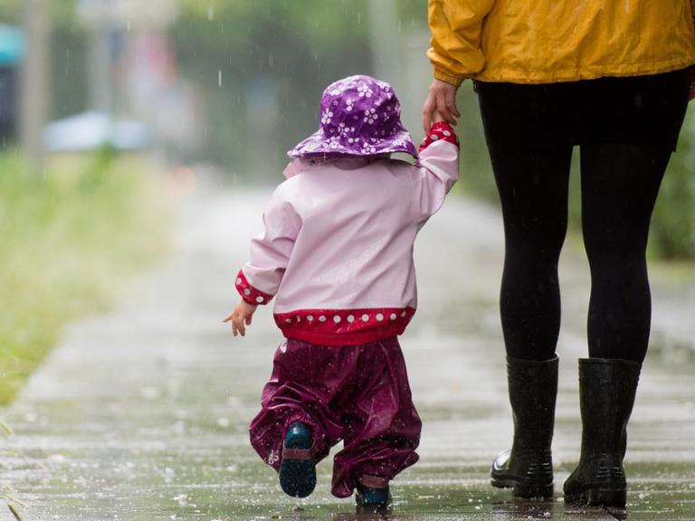 Ein kleines Mädchen läuft mit Regenkleidung an der Hand ihrer Mutter über einen Gehweg