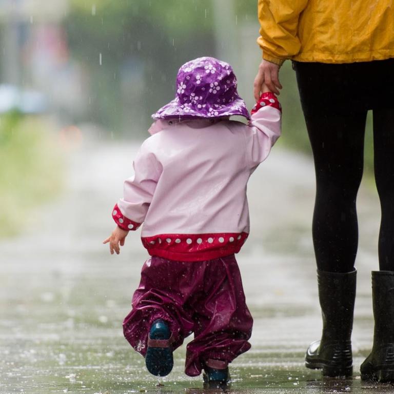 Ein kleines Mädchen läuft mit Regenkleidung an der Hand ihrer Mutter über einen Gehweg