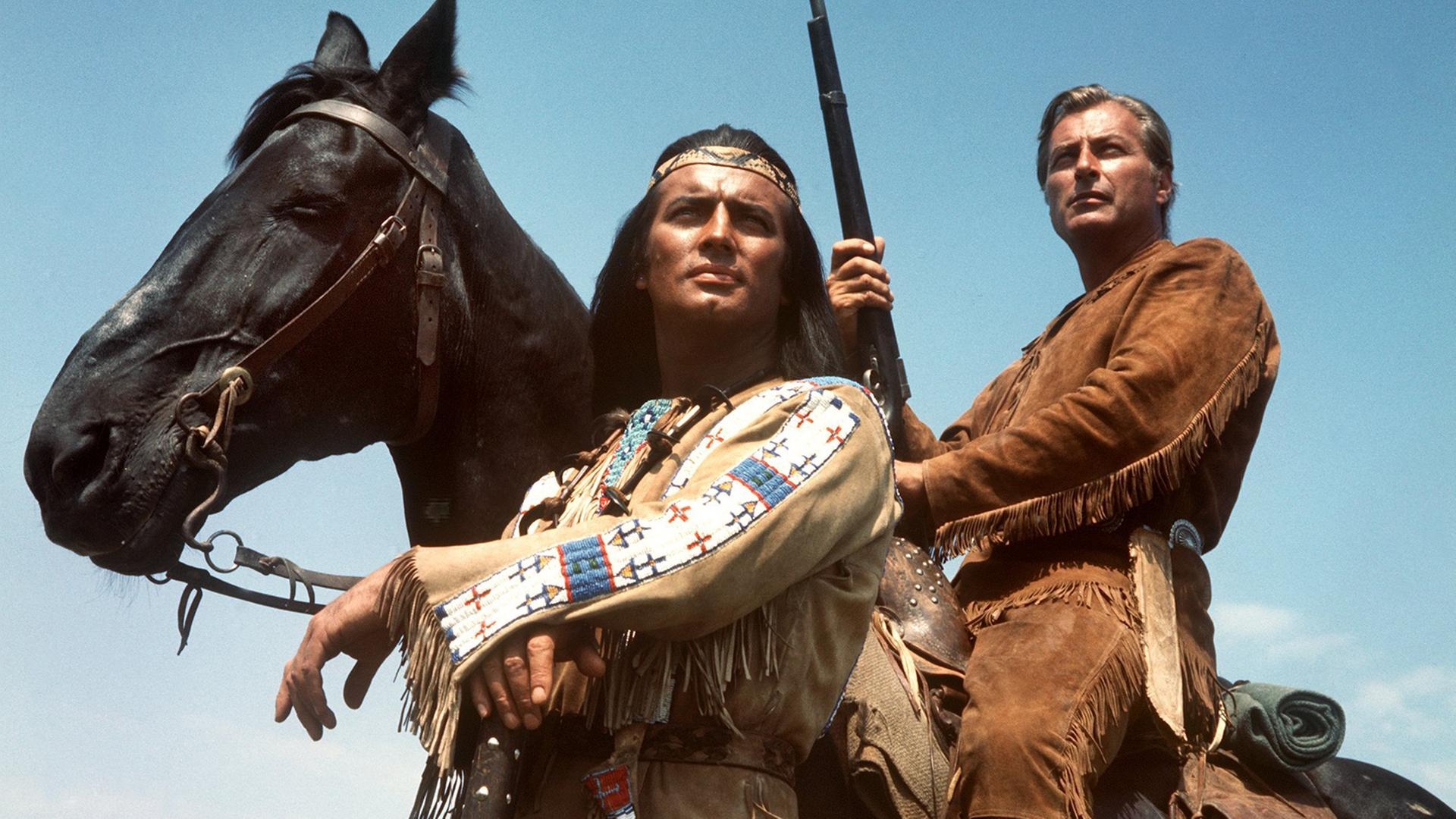Pierre Brice (l) als Apachen-Häuptling Winnetou und Lex Barker als sein Blutsbruder Old Shatterhand in einer Szene des Karl-May-Films "Im Tal des Todes". Lex Barker sitzt auf einem Pferd mit einem Gewehr in der Hand. Beide schauen in die Ferne.