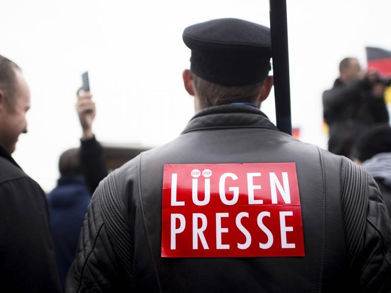Ein Mann ist von hinten zu sehen. Er trägt eine Schiebermütze und auf seinem Rücken steht "Lügenpresse". Dahinter ist eine Deutschlandfahne zu sehen.