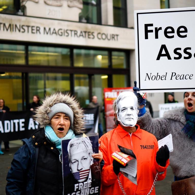 Im Vordergund demonstrieren drei Menschen mit Pappschildern und Masken für die Freilassung von Julian Assange. Im Hintergrund ist der Eingang vdes Westminster Magistrates Courts  zu sehen.