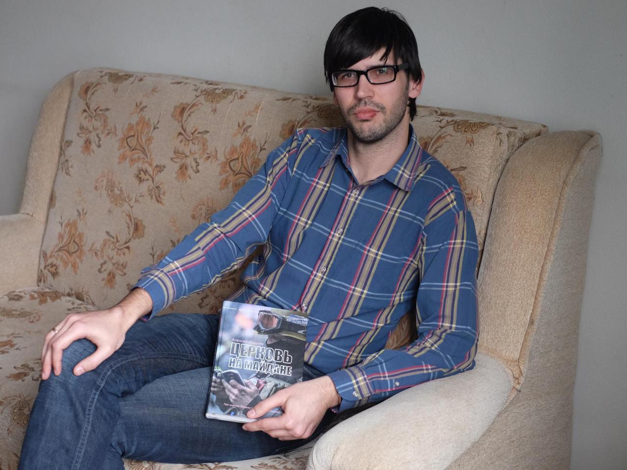 Der Buchautor Alexej Gordjejew sitzt auf einer Couch und hält sein Buch über die Rolle der Kirche auf dem Maidan in den Händen. 