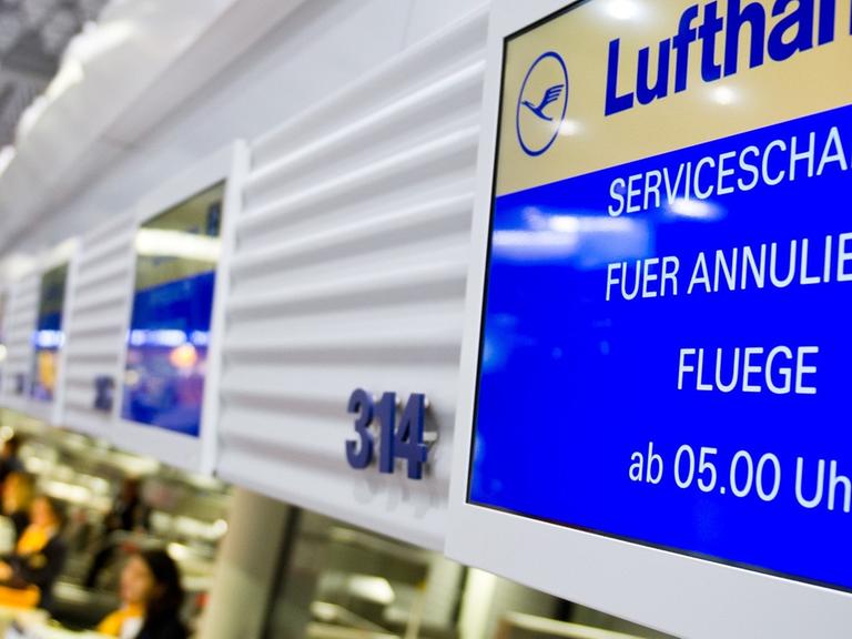 An einem Flugschalter der Lufthansa in Frankfurt am Main ist auf dem Display zu lesen: "Serviceschalter für annulierte Flüge ab 5 Uhr." Vor dem Schalter sind unscharf Fluggäste zu erkennen; dahinter sitzen zwei Service-Angestellte der Lufthansa.