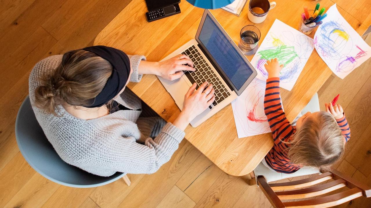 Ein zweijähriges Kind malt ein Bild, während seine Mutter im Homeoffice an einem Laptop arbeitet.