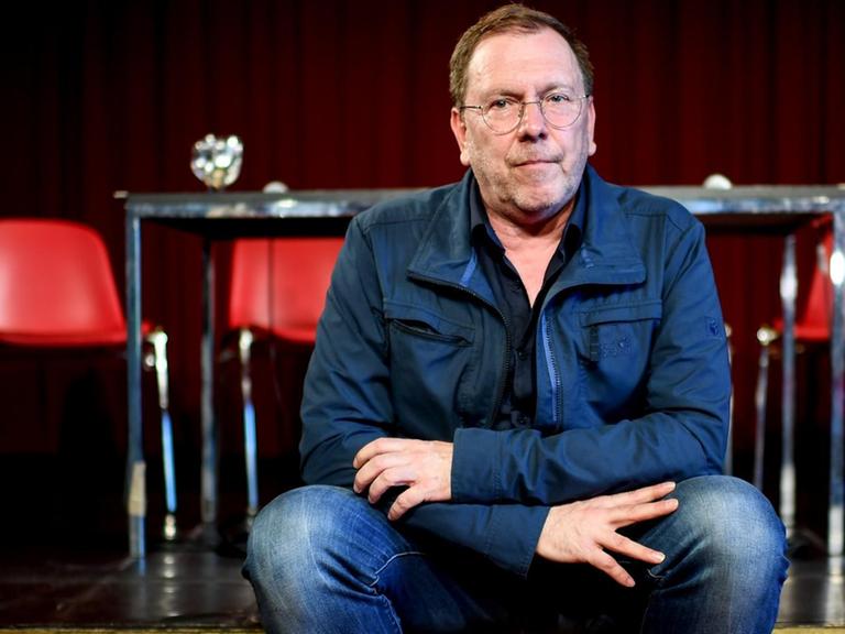 René Pollesch, Theaterregisseur und Autor, sitzt am Rande einer Pressekonferenz zur "Zukunft der Volksbühne". Er sitzt auf einer niedrigen Bühne, im Hintergrund sind ein Tisch und vier rote Stühle zu sehen, 2019.