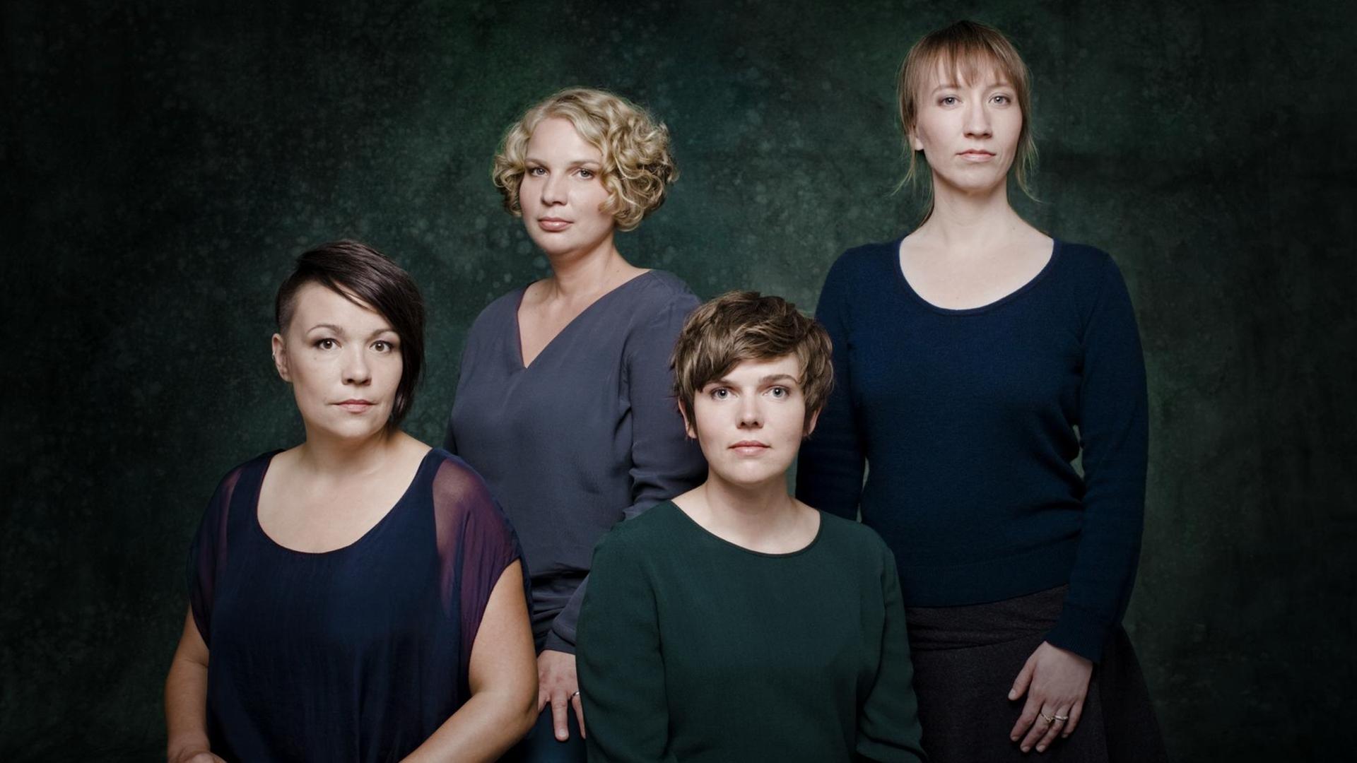 Vier Frauen unterschiedlichen Types stehen dicht beieinander und schauen ernst in die Kamera.