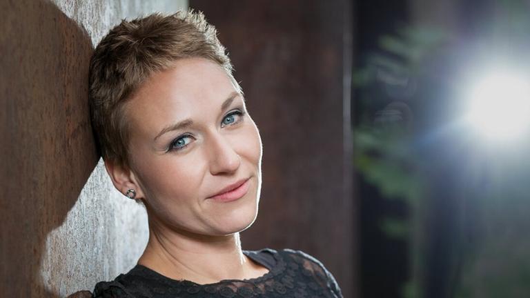 Eine junge Frau mit sehr kurzem Haar lehnt in einem Spitzentop an einer Wand und lächelt in die Kamera. Eine junge Frau mit sehr kurzem Haar lehnt in einem Spitzentop an einer Wand und lächelt in die Kamera.