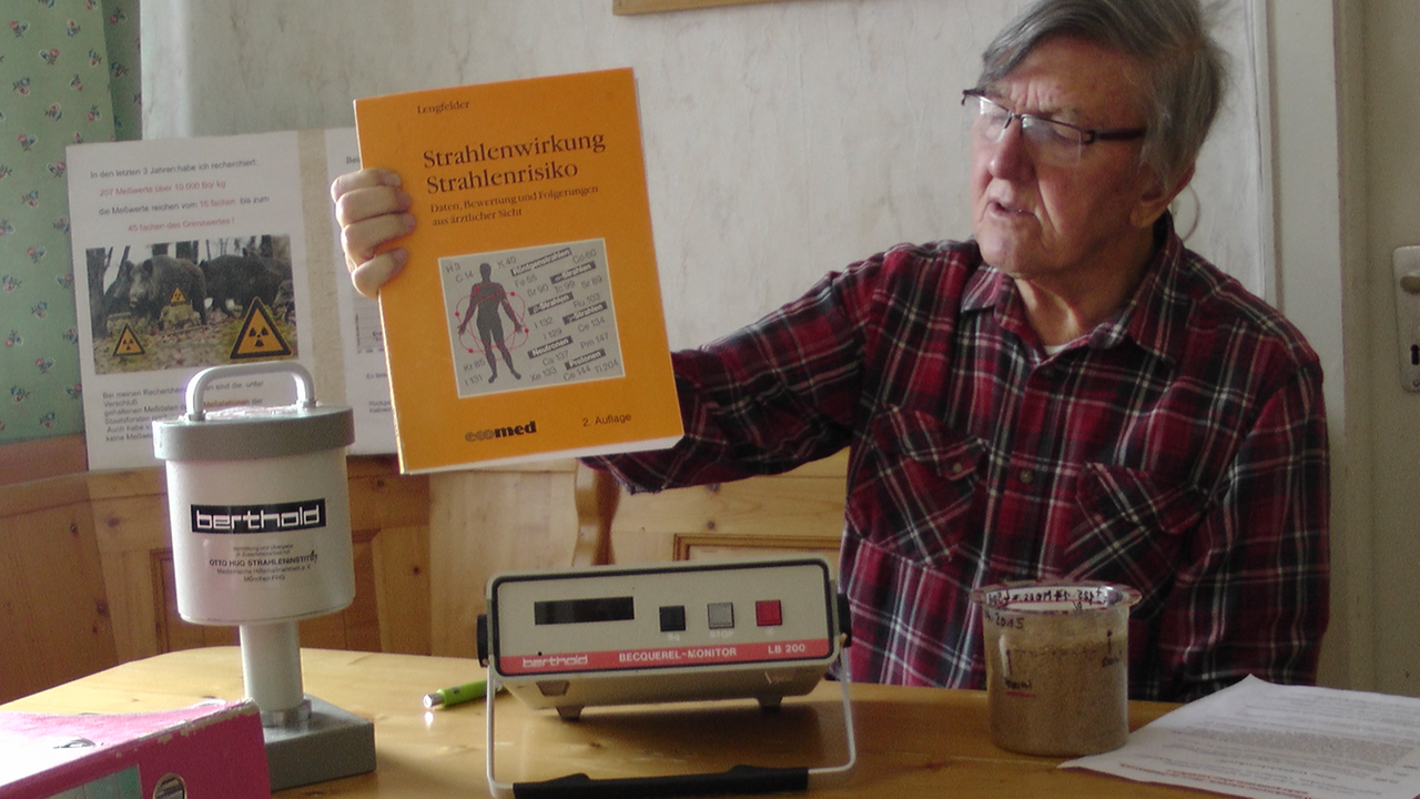 Helmut Rummel sitzt an seinem Küchentisch. In der Hand hält er ein Buch über Strahlenrisiken. Auf dem Tisch steht ein Geigerzähler. 