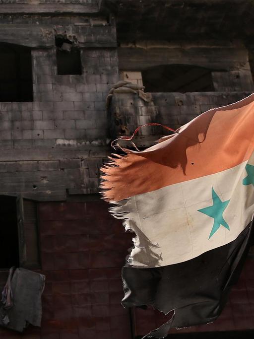 Eine an den Rändern zerfetzte syrische Fahne wehr vor einem zerstörten Gebäude in Homs