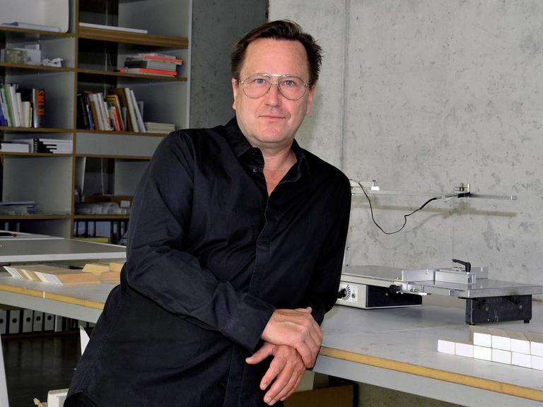 Arno Brandlhuber steht, mit dem Rücken an einen Arbeitstisch gelehnt, in seinem Studio. Im Hintergrund ist eine Sichtbeton-Wand und ein Bücherregal zu sehen.