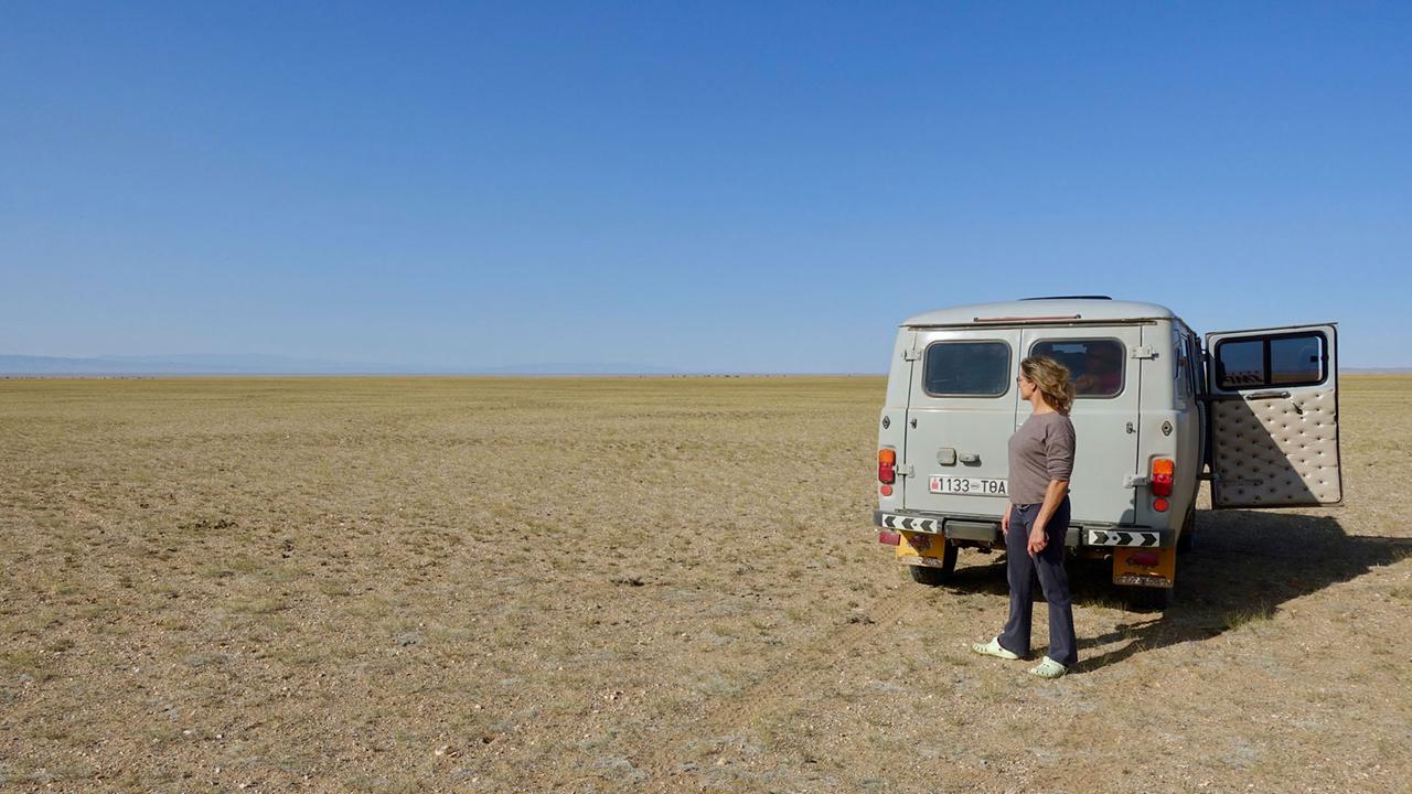 In der Wüste Gobi steht ein VW-Bus und eine Frau davor. Die Sonne scheint und der Himmel ist blau. 