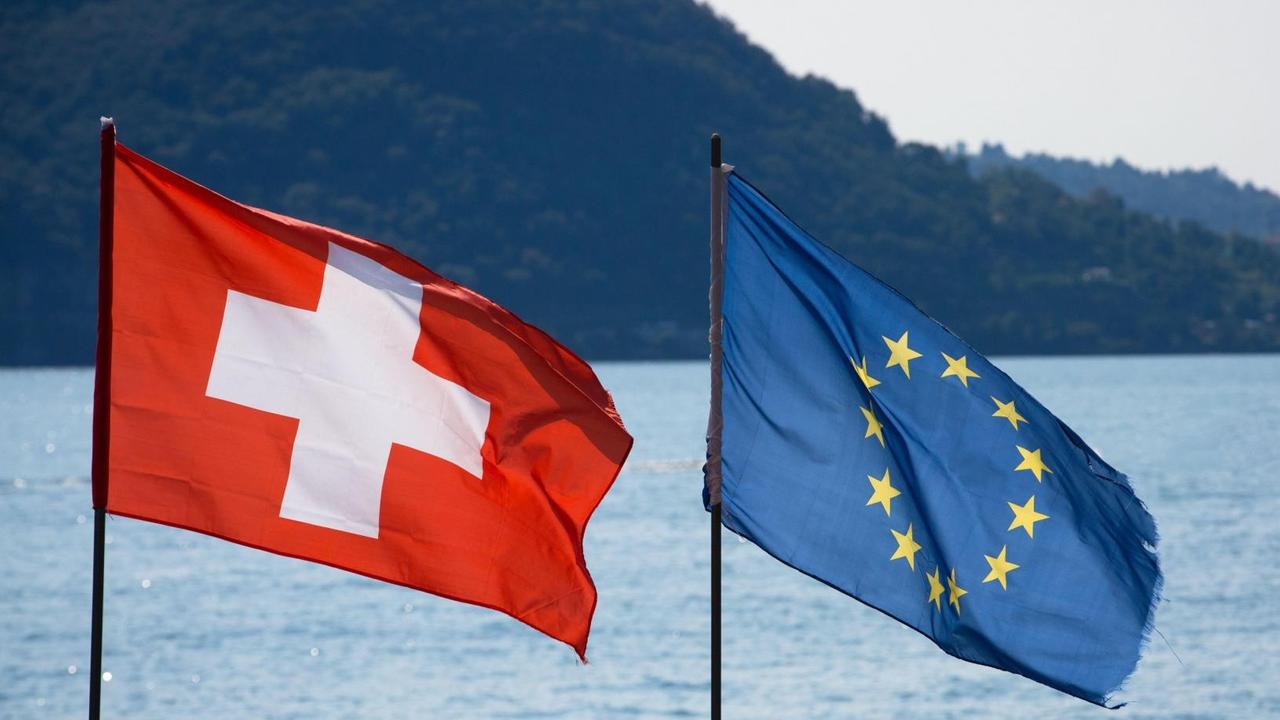 Ein Farbfoto zeigt die Flaggen der Europäischen Union und der Schweiz nebeneinander am Ufer eines Bergsees