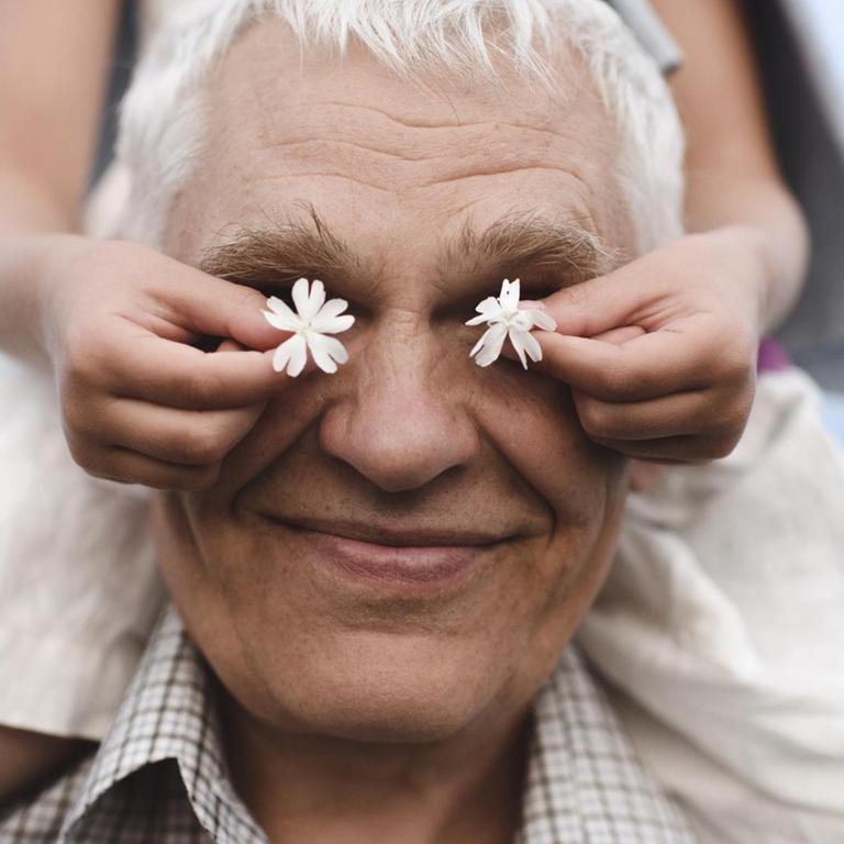 Ein lachender Großvater trägt ein Mädchen auf den Schultern. Es hält ihm zwei weiße Blumen vor die Augen.