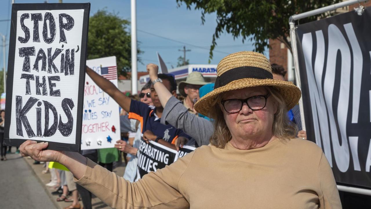 Die ältere Frau mit Strohhut hält das Schild und blickt in die Kamera. Dahinter sieht man weitere Demonstranten mit Schildern am Straßenrand.