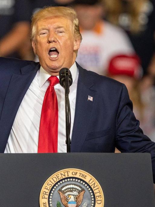 US-Präsident Trump steht an einem Rednerpult und gestikuliert mit der rechten Hand. Im Hintergrund sind Zuhörer zu sehen.