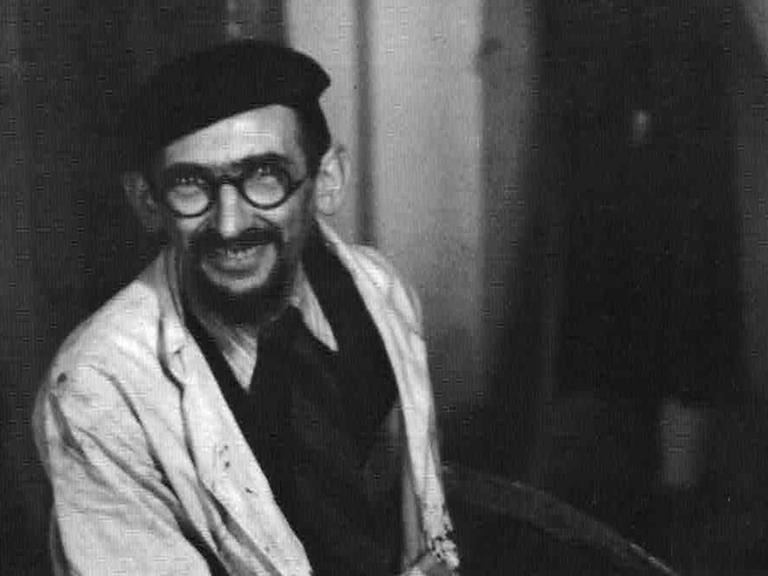 Schwarzweißfoto des Malers Horst Strempel, der in einem Kittel in seinem Atelier sitzt und freundlich in die Kamera blickt.