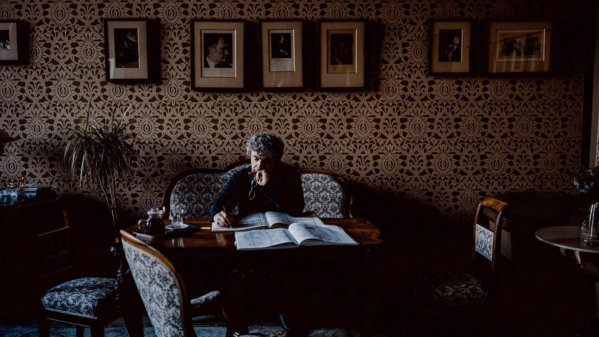 Semyon Bychkov sitzt auf einer antiken Couch, vor sich ein Tisch mit einer Partitur, in die er Notizen schreibt