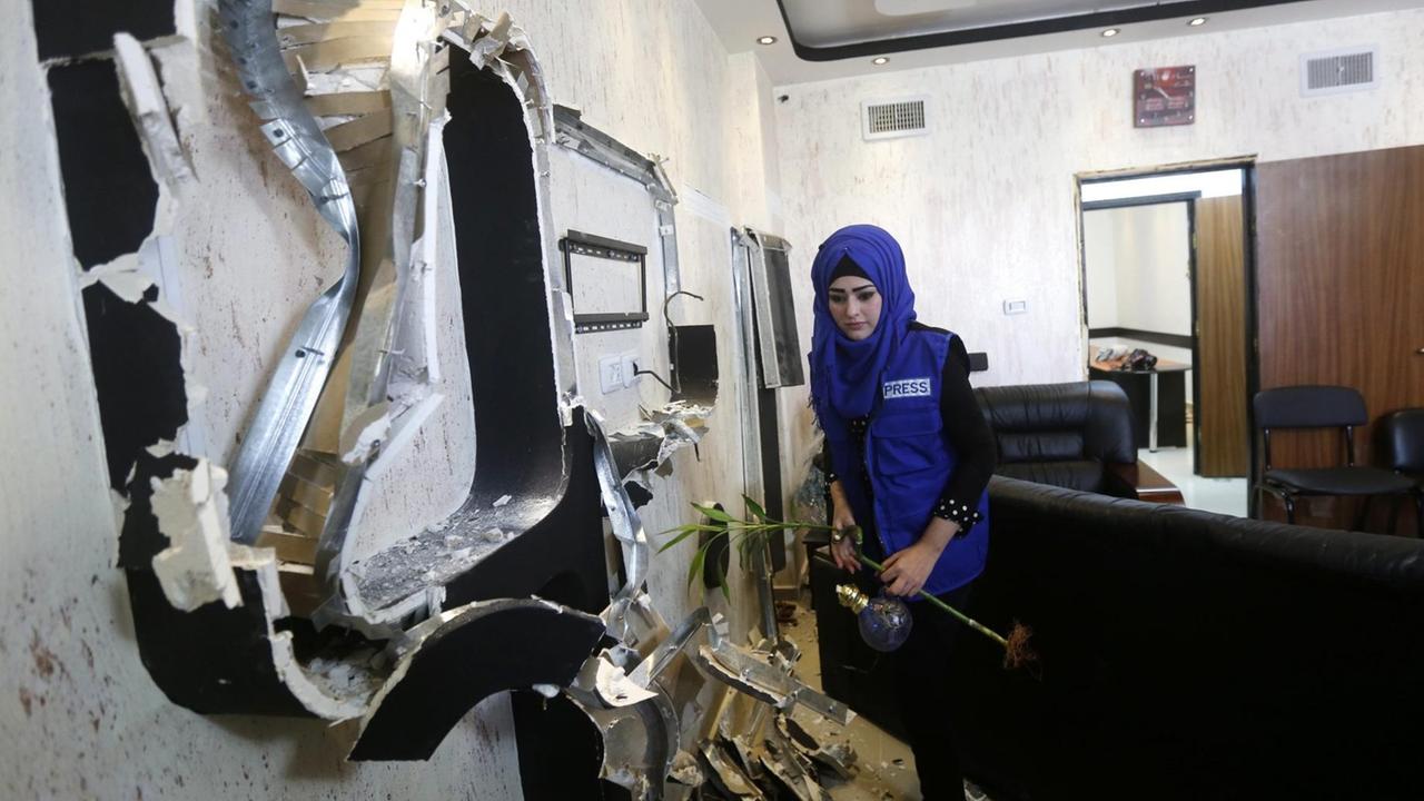 Die Journalistin trägt ein blaues Kopftuch und eine blaue Weste mit der Aufschrift "Press". Sie betachtet in einem Büro eine Wand, aus der Monitore und Kabel herausgerissen wurden. In der Hand hält sie eine Vase und eine Pflanze. Die Journalistin trägt ein blaues Kopftuch und eine blaue Weste mit der Aufschrift "Press". Sie betachtet in einem Büro eine Wand, aus der Monitore und Kabel herausgerissen wurden. In der Hand hält sie eine Vase und eine Pflanze.
