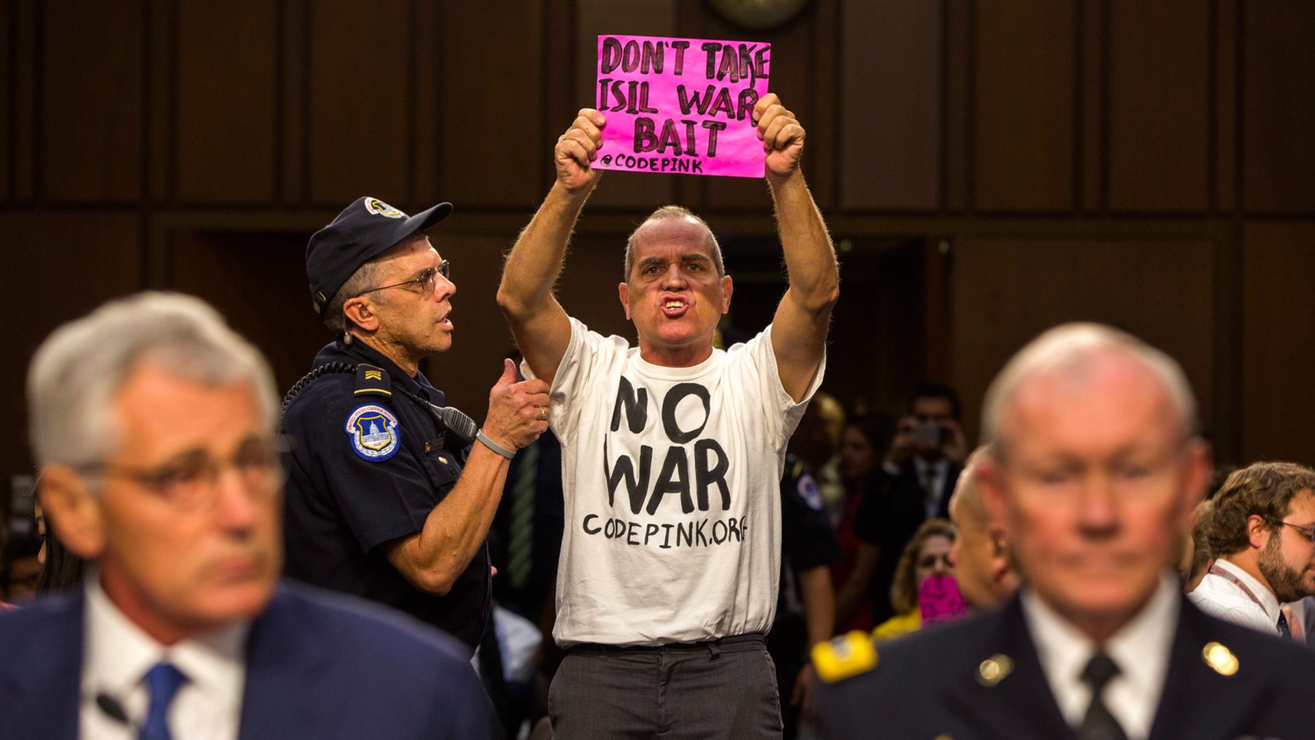 Chuck Hagel und Martin Dempsey sitzen an einem Tisch, während hinter ihnen der Aktivist Tighe Berry aufsteht. Dieser trägt ein T-Shirt mit der Aufschrift "No War" und ein pinkfarbenes Schild mit dem Text "Don´t take Isil war bait"
