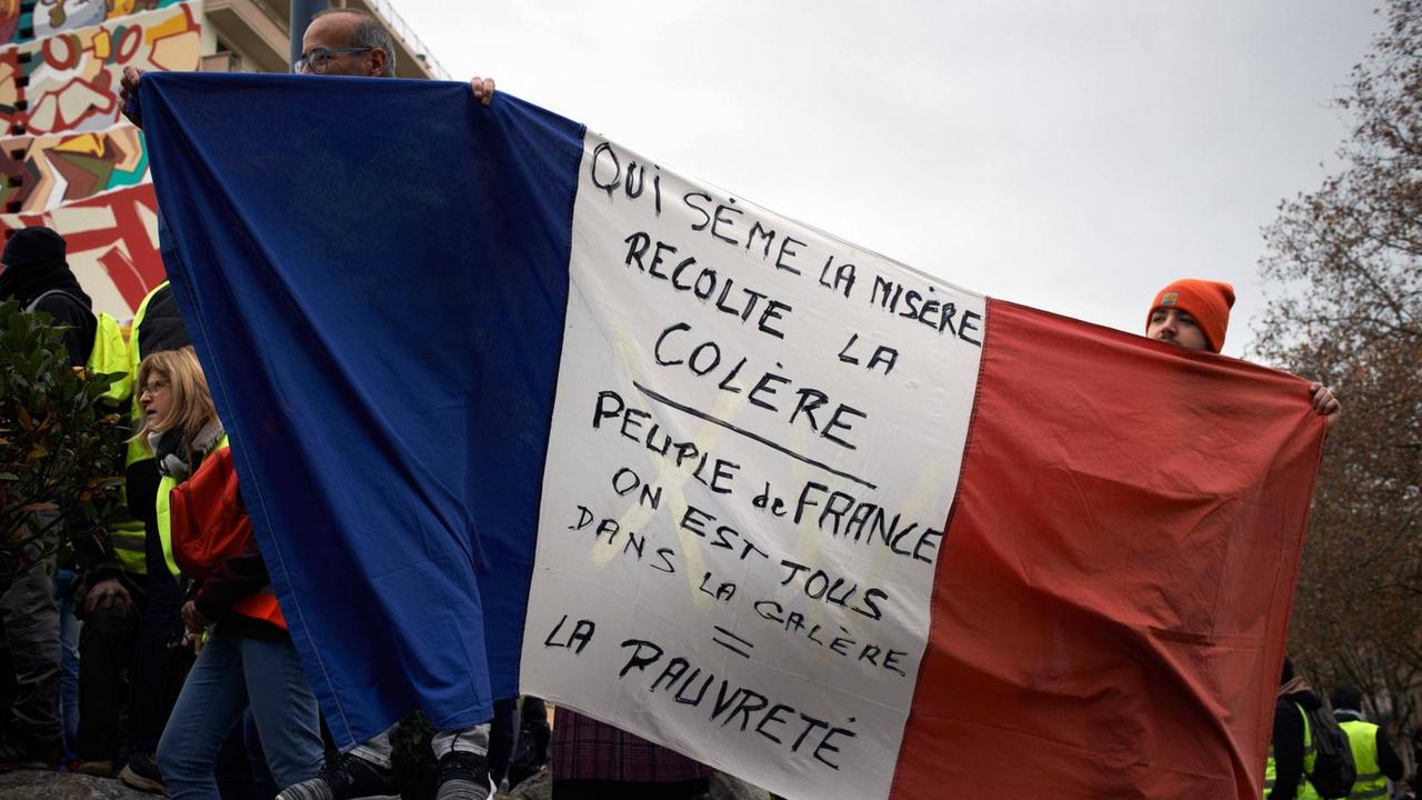 Menschen in Frankreich protestieren gegen die Armut im Land. Sie halten die französische Nationalflagge hoch auf der geschrieben steht: "Wer die Armut sät, erntet die Wut."