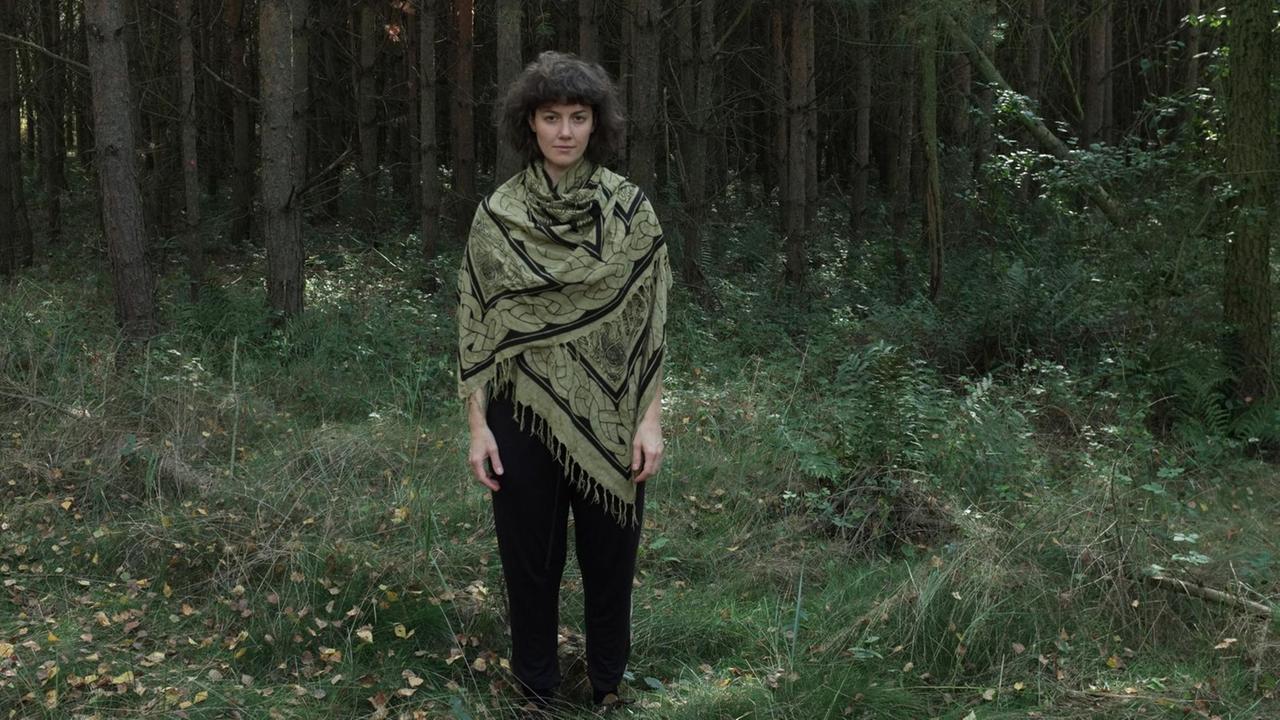 Eine junge dunkelhaarige Frau steht mitten einem Wald. Sie trägt ein großes Tuch um die Schultern gewickelt.