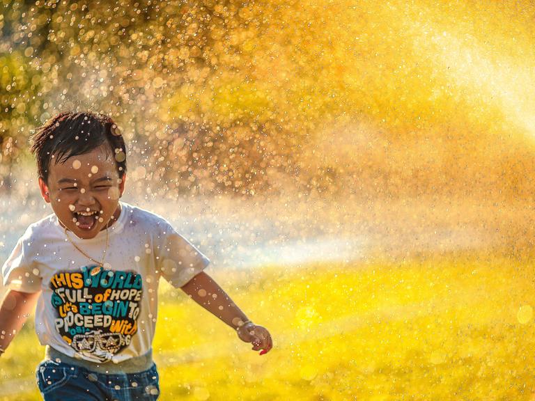 Ein kleiner Junge rennt begeistert unter dem Wassernebel einer Sprenkleranlage her